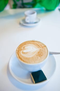 Closeup of espresso and milk based latte in cup with heart shape coffee art design and wrapped chocolate with espresso cup in background