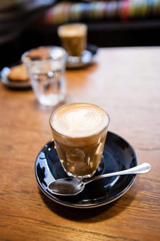 Closeup of espresso and milk based latte in glass with heart shape coffee art design resting on saucer on wooden table