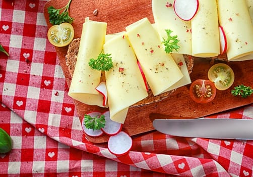 Gouda cheese rolls on bread seasoned with pepper and topped with radish, parsley and tomato served on a red and white checked country tablecloth, high angle view