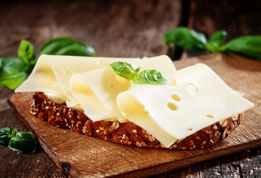 Thinly sliced gouda cheese on wholewheat bread garnished with fresh basil served on a wooden board, close up view
