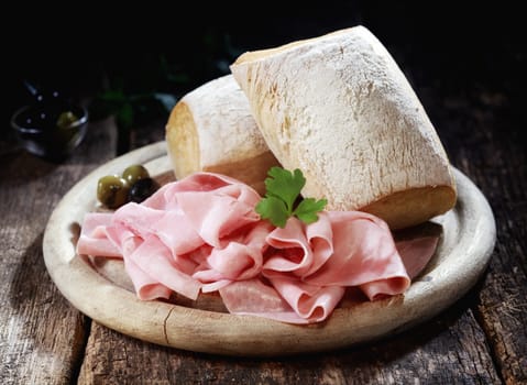 Country lunch with thinly sliced cured Bavarian ham and crusty rolls served on a wooden platter on a rustic wooden table