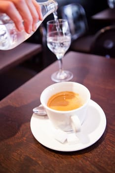 Closeup of cup of espresso coffee on wooden cafe table and pouring of water into glass from bottle in background