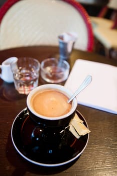 Closeup of cup of espresso coffee with metal spoon on wooden cafe table with glass of water and small jug and ashtray in background