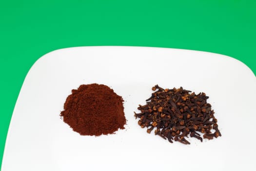 A half ounce pile of cloves next to a half ounce of whole clove buds on a white plate with a green background..