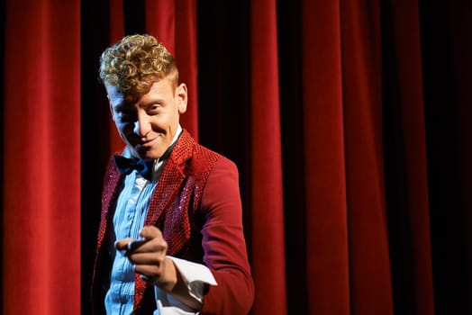 Arts and entertainment in theatre with funny man working as anchorman, standing against red curtains and pointing to camera
