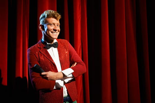 Arts and entertainment in theatre with funny man working as anchorman, standing with arms crossed against red curtains with microphone