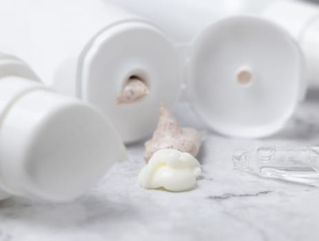 White tube and bottle with open cap and beige cream and scrub squeezed on white marble table, against cosmetic bottles, close up. Natural product for daily face care, spa procedures