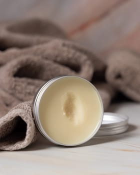 Opened body butter jar near light brown bath towel in bathroom, close up. Natural herbal cosmetic product for body care. Daily beauty routine or spa procedures
