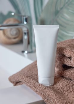 White cream squeeze tube on brown folded towels on basin in green tropical bathroom close up, packaging mockup. Natural plant unbranded cosmetics with copy space. Spa and wellness concept