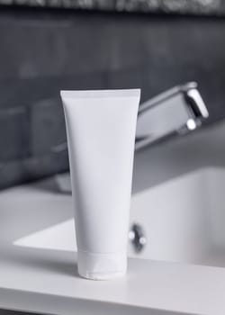 White cream squeeze tube near modern faucet on basin in dark grey masculine bathroom close up, packaging mockup. Natural unbranded cosmetics with copy space. Spa and wellness concept