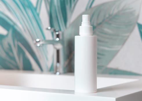 White cosmetic spray bottle on washbasin with faucet in green tropical bathroom close up, packaging mockup. Natural plant unbranded lotion or toner container, copy space. Spa and wellness concept