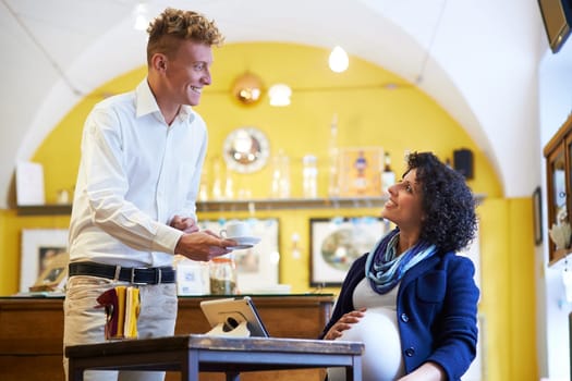 People in cafeteria, with barman serving espresso coffee to pregnant businesswoman sitting at table with ipad