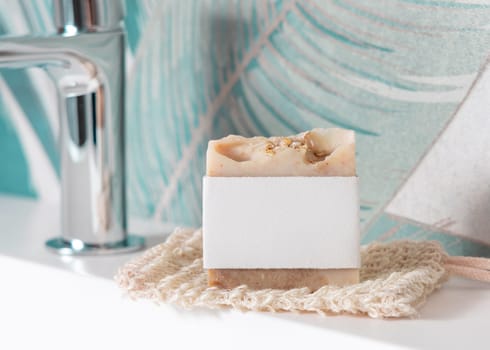 Beige handmade soap bar with blank label on sisal soap saver bag on washbasin near faucet in green tropical bathroom close up, mockup, copy space. Natural herbal unbranded product for daily hygiene