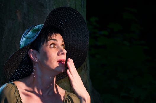 Side head-and-shoulder portrait of a middle-aged black-haired woman with a dark sun hat on a tree in the narrow beam of light looking up towards the light
