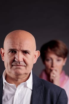 an old man in foreground and an old woman in background looking at him with concerns about him
