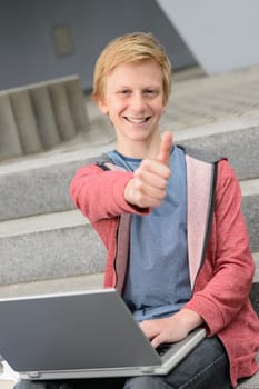 Successful teenage student show thumb-up  sitting with laptop on steps