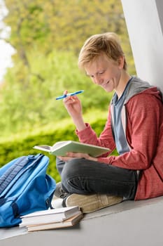 Smiling teenage boy reading book sitting on wall outside school