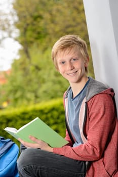 Happy teenage boy with book sitting outside on wall
