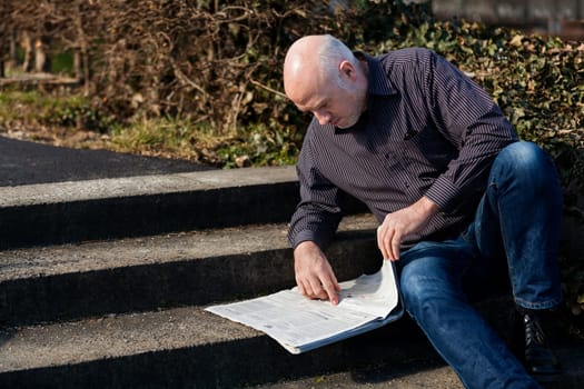 Middle-aged man,senior sitting with a newspaper on a staircase in spring outdoors