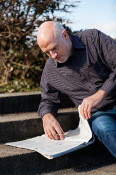 Middle-aged man,senior sitting with a newspaper on a staircase in spring outdoors