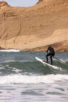 One man paddles out into the ocean on long paddle board near rock formation