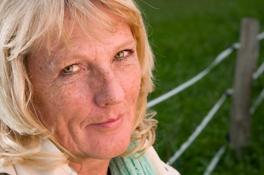 Side close-up of a blonde mature woman on a green meadow at a fence friendly looking into the camera