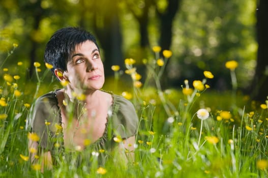 frontal head-and-shoulder view of a middle-aged black-haired woman on her belly in the grass with yellow flowers looking dreamily up the right