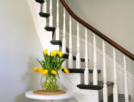 Yellow tulips in a round glass bowl on a white round table beside heritage staircase with dark stairs and white spindles