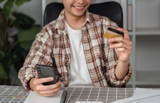 A young man analyzes his expenses using a smartphone and credit card, feeling a sense of accomplishment and joy.