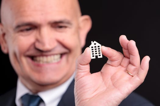 an old self confident man smiling with a little wooden house between his fingers