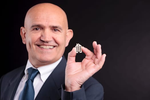 an old self confident man smiling with a little wooden house between his fingers