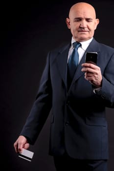 an old businessman with a credit card and a mobile hphone making purchases online  DARK BACKGROUND