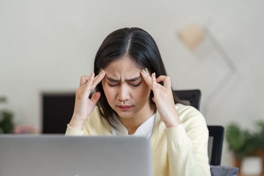 A focused Asian woman holds her temples in discomfort while working on a laptop, representing stress and mental health in the workplace.