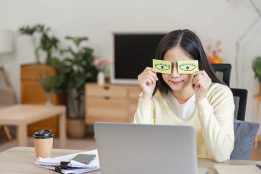 A cheerful Asian woman playfully holds up paper eyes in front of her face while working on a laptop, highlighting creativity and a lighthearted approach to remote work.