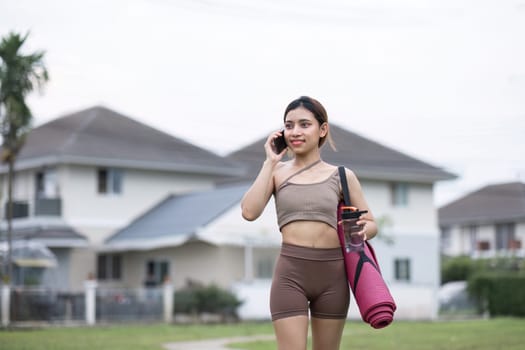 Young healthy woman holds an exercise mat. Use your smartphone while waiting to do outdoor yoga on the grass at the park..