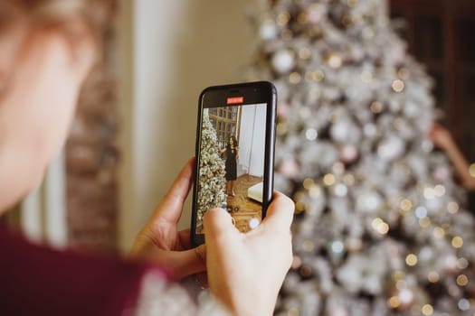 Girl's hand holding mobile phone take a photo of christmas tree .Young woman using smartphone outdoor capture picture of bokeh blur Christmas lighting lamp decoration.