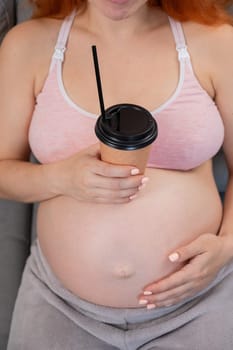 Faceless pregnant woman in 3rd trimester holding a coffee glass on her belly