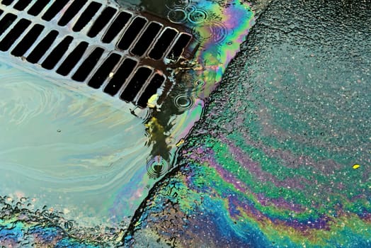 An oil slick against the backdrop of an asphalt road flows into a storm drain through a grate.