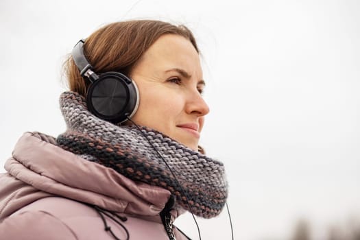 Joyful young woman in jacket listening to music via black headphones, breathing in the fresh air, relaxing in a park