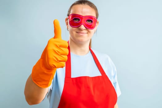 Young confident woman in red mask, gloves and apron showing ok sign with thumb up on blue background. Super housewife