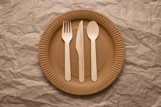 Wooden fork, spoon and knife on cardboard plate on crumpled paper background. Biodegradable eco-friendly dishes. Top view