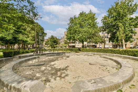 a sand pit in the middle of a park with trees and buildings in the background on a bright sunny day