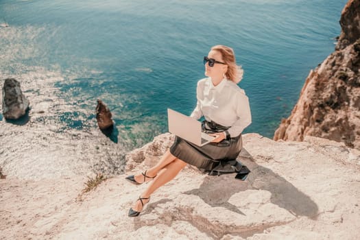 Business woman on nature in white shirt and black skirt. She works with an iPad in the open air with a beautiful view of the sea. The concept of remote work