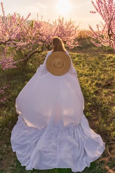 Woman blooming peach orchard. Against the backdrop of a picturesque peach orchard, a woman in a long white dress and hat enjoys a peaceful walk in the park, surrounded by the beauty of nature