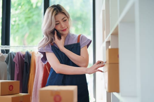 Starting small business entrepreneur of independent Asian female online seller talking on the phone with a customer and packing products for delivery to the customer. and SME delivery concept.