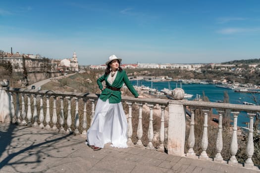 Woman walks around the city, lifestyle. Happy woman in a green jacket, white skirt and hat is sitting on a white fence with balusters overlooking the sea bay and the city