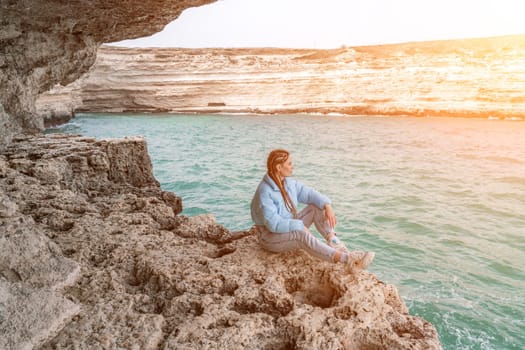 woman sea travel. A woman in a blue jacket sits on a rock above a cliff above the sea, looking at the stormy ocean. Girl traveler rests, thinks, dreams, enjoys nature.