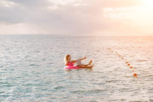 Woman works on laptop in sea. Freelancer, blond woman in sunglases floating on an inflatable big pink donut with a laptop in the sea at sunset. People summer vacation rest lifestyle concept