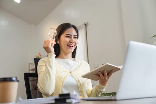 A joyful young woman holds a light bulb in one hand while engaging with her laptop, symbolizing creativity and inspiration in her workspace.