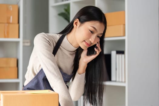 A focused young woman in an apron talks on the phone while reviewing paperwork and managing inventory in a well-organized workspace.